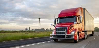 Red commercial semi-truck driving down a highway alongside open grassy fields.